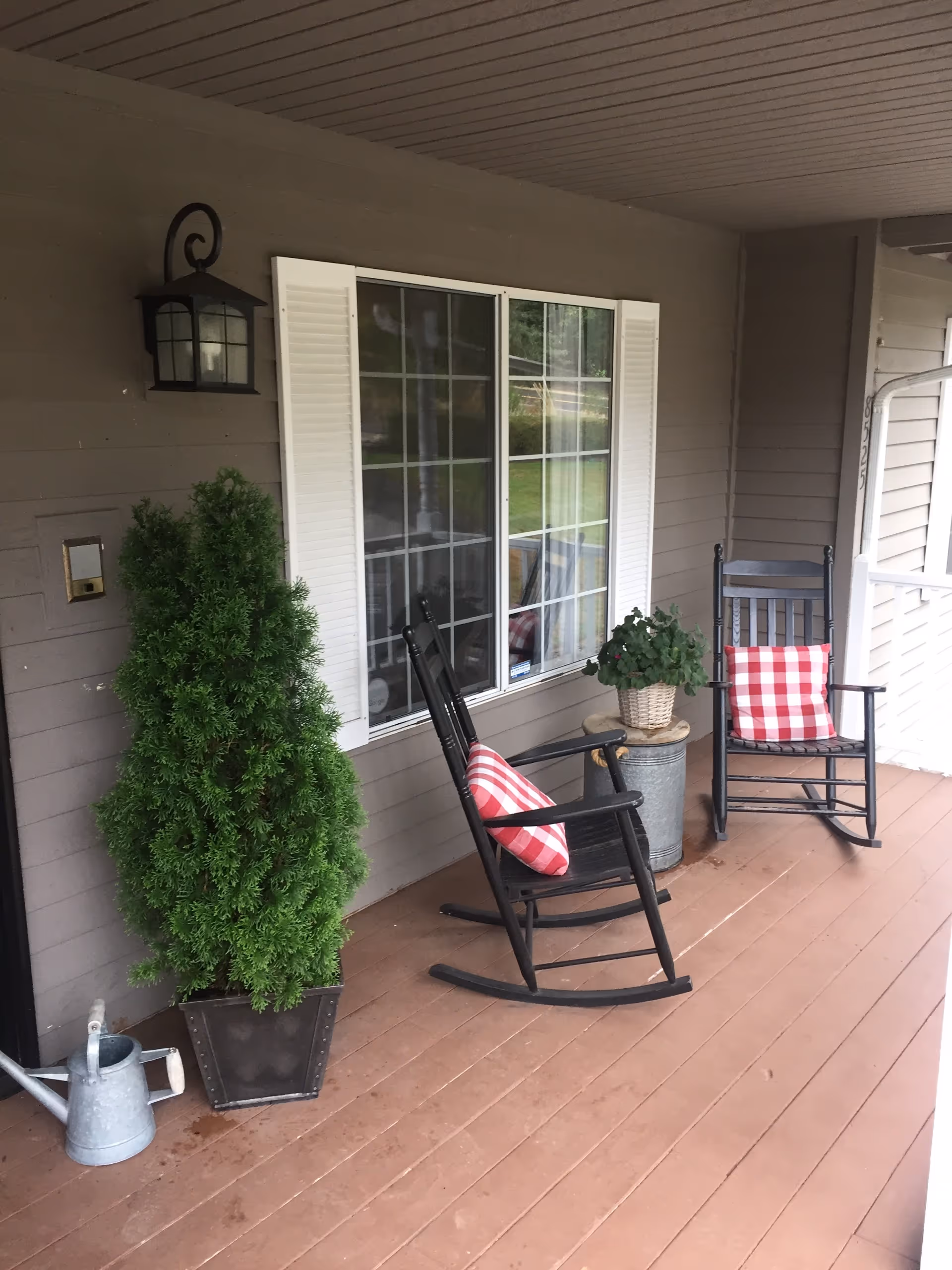 A covered porch area with two black rocking chairs, each with a red and white checkered cushion. There is a potted green plant in a metal container between the chairs, a tall green potted shrub on the left side, and a metal watering can on the floor. The porch has a wooden floor and beige siding with a window featuring white shutters.