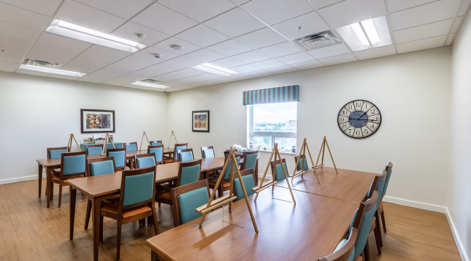 Well-lit activity/dining room with rows of wooden tables, teal-upholstered chairs, and small easels on the tables.