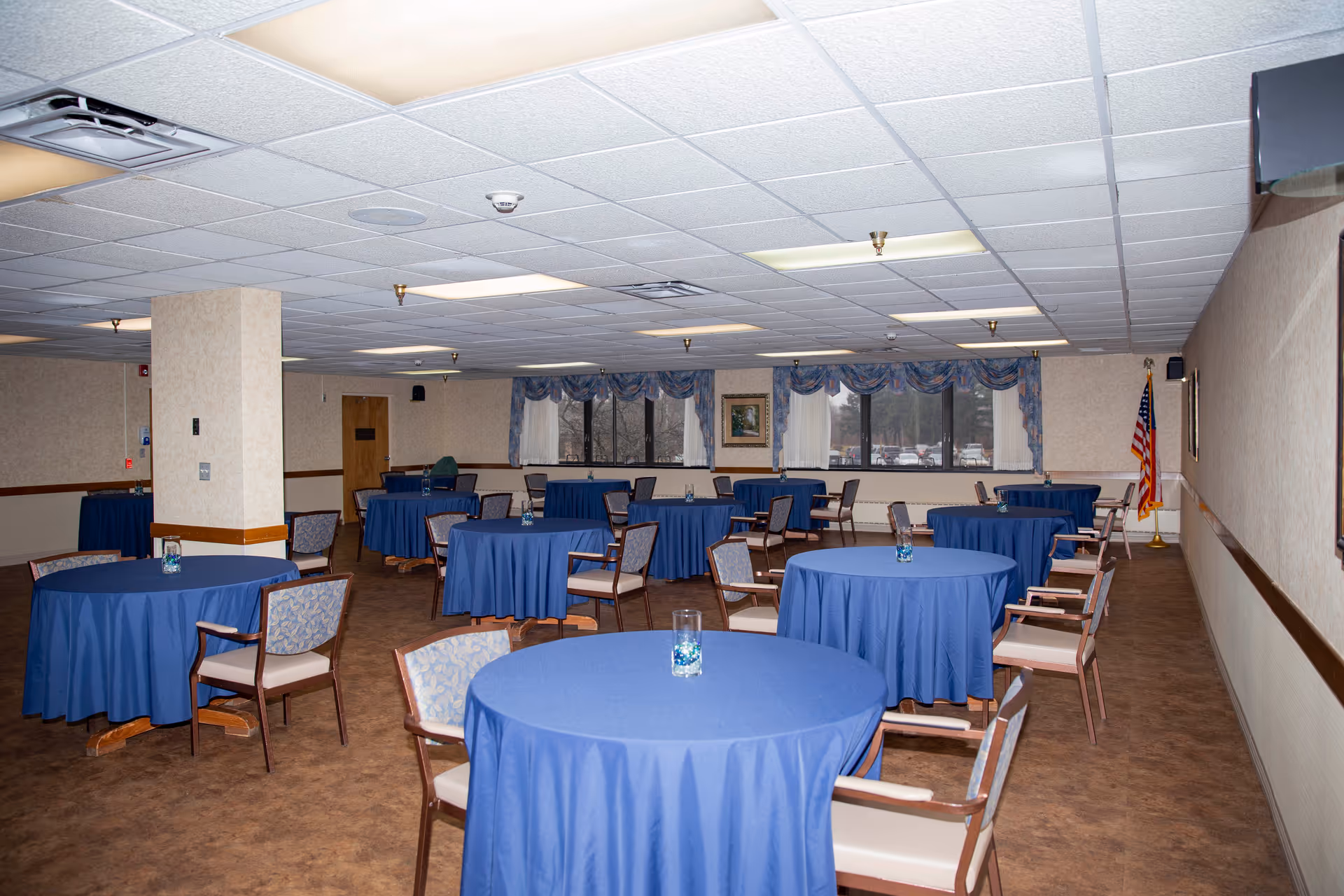 A large dining room with multiple round tables covered in blue tablecloths, each surrounded by chairs. The room has a drop ceiling with fluorescent lighting, windows with blue valances, and an American flag in the corner. The floor is carpeted and the walls are beige with a wooden trim.