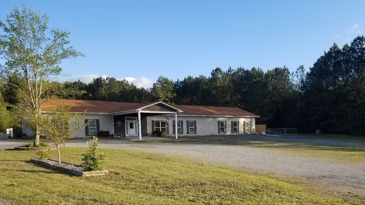 Single-story light-colored care home with a red roof and front porch set on a grassy lot with trees in the background.