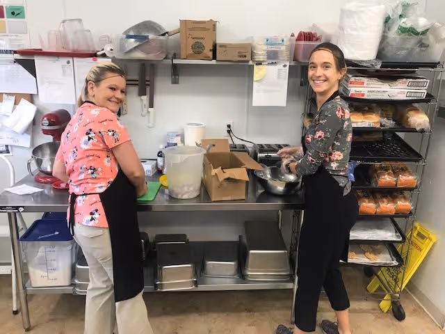 Two women wearing aprons are smiling and preparing food in a commercial kitchen area with stainless steel counters, shelves stocked with bread and supplies, and various kitchen utensils.