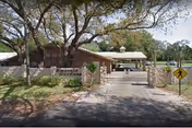 Entrance to Emerald Gardens facility showing a gated driveway with a stone wall and sign reading Emerald Gardens. Large trees provide shade over the entrance area.