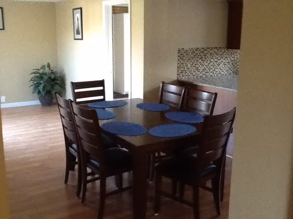 A dining area with a dark wooden rectangular table surrounded by eight matching chairs. The table has six blue round placemats. The room has wooden flooring, beige walls, a potted plant in the corner, and a small tiled backsplash area visible in the background.