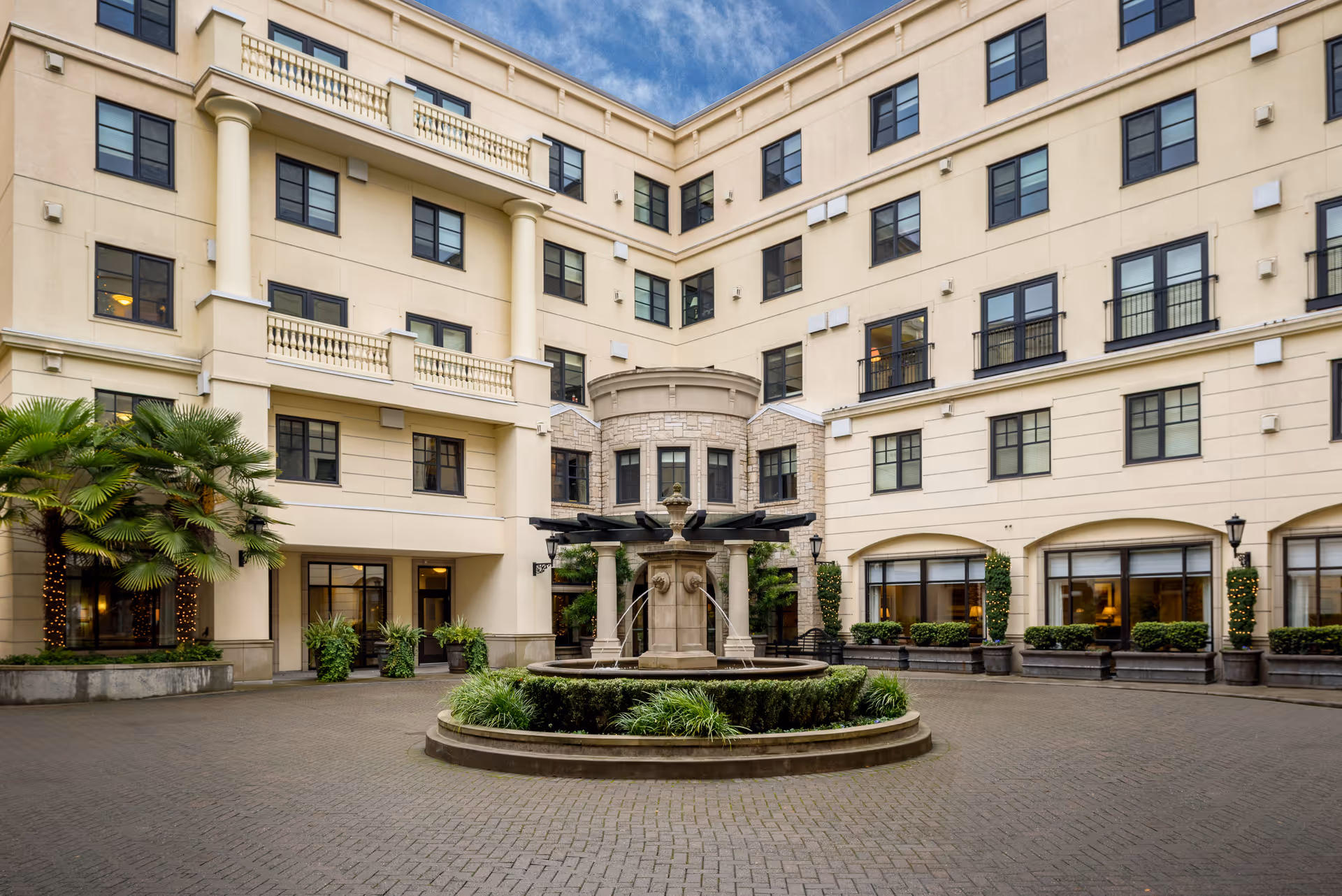 A landscaped interior courtyard of a multi-story beige building featuring a circular fountain, potted plants, palm trees, and many windows.