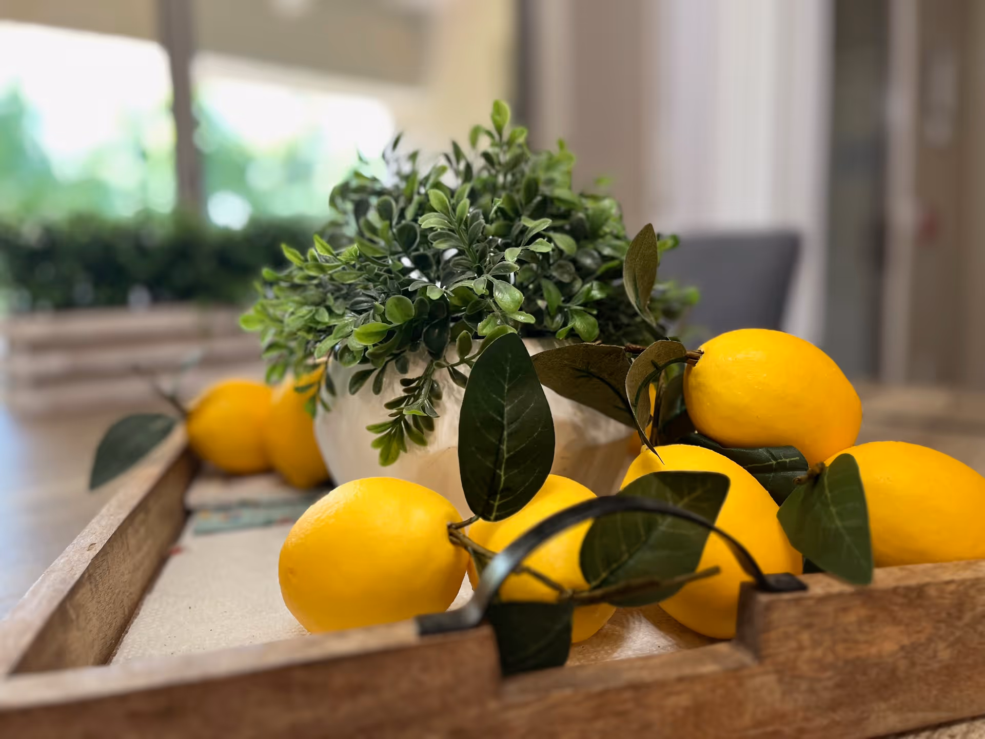 A wooden tray holding a white pot with green leafy plant and several bright yellow lemons with green leaves, placed on a table in a softly lit room with blurred background.