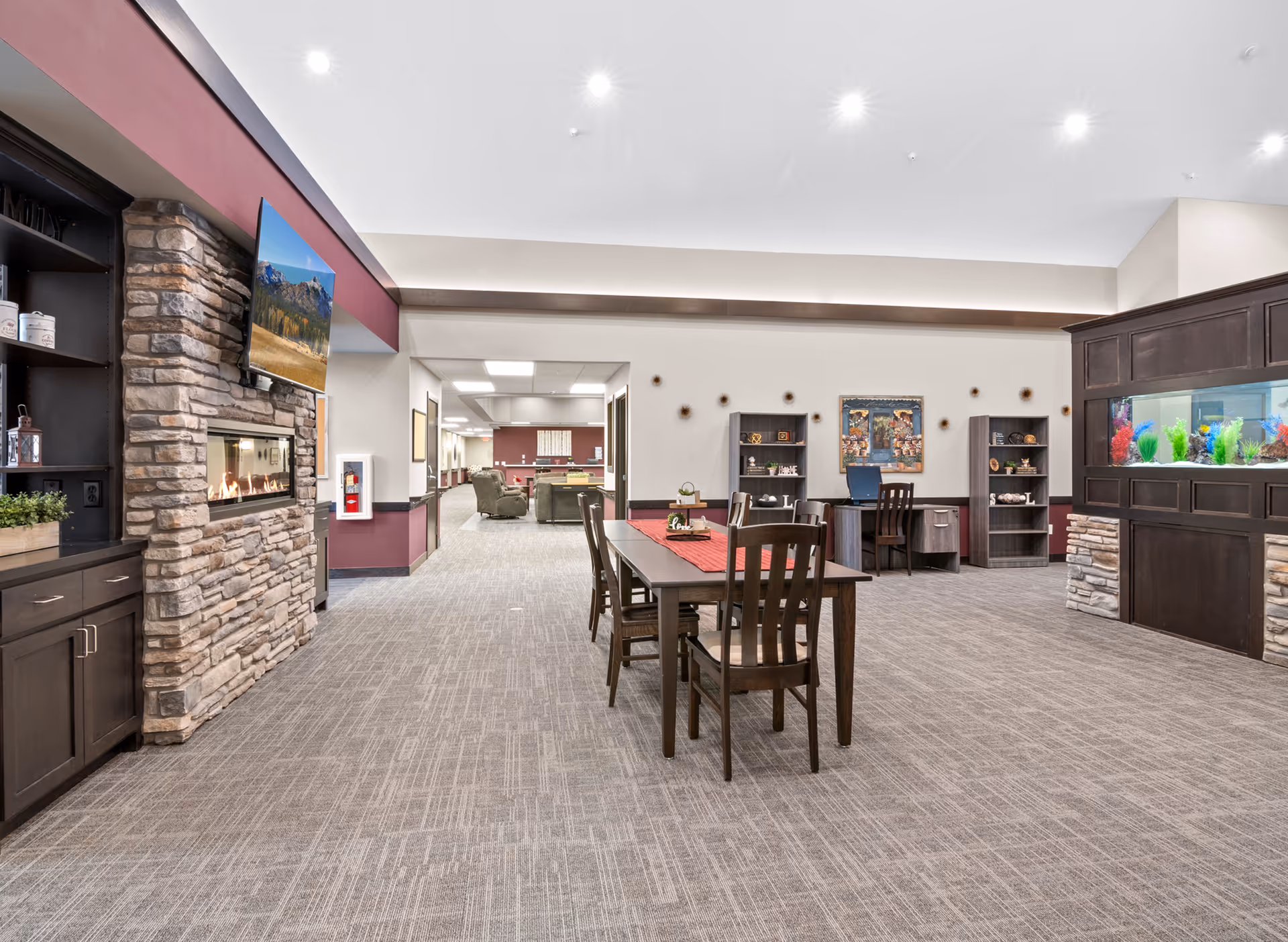 A spacious and well-lit common area in a senior living facility featuring a stone fireplace with a mounted TV above it on the left, a long wooden table with chairs in the center, and a large built-in aquarium on the right. The room has carpeted floors, neutral walls with maroon accents, and decorative shelves and artwork on the far wall. Comfortable seating areas are visible further back in the room.