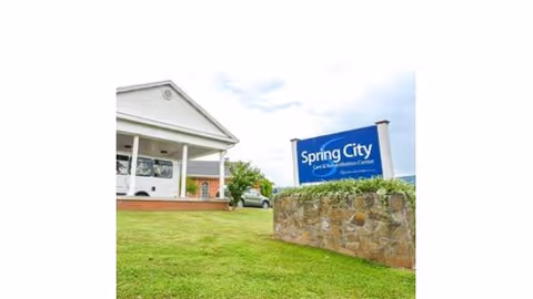 Exterior view of Spring City Care and Rehabilitation Center showing a white building with a covered entrance and a blue sign with the facility's name next to a stone wall on a grassy lawn under a partly cloudy sky.