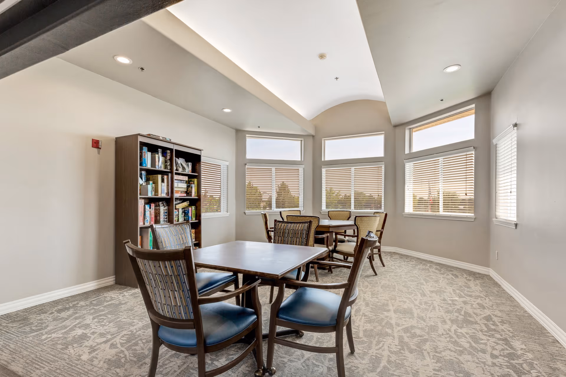 A bright room with large windows featuring blinds, several wooden tables with chairs around them, and a bookshelf filled with board games and books. The room has a patterned carpet and recessed ceiling lights.
