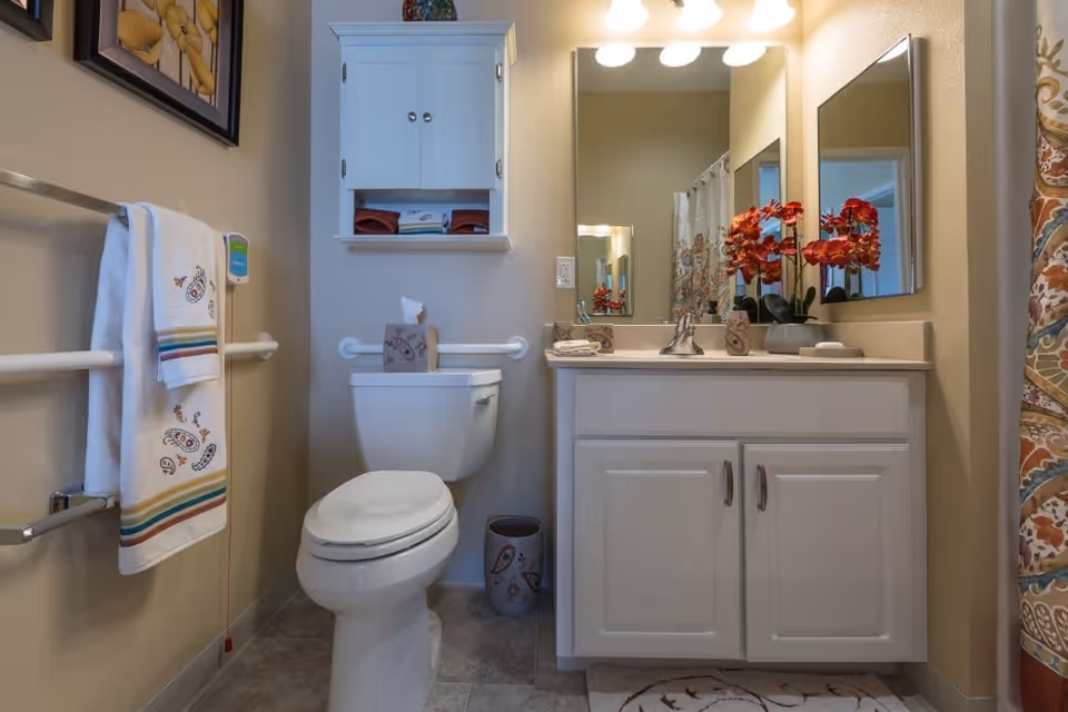 A clean and well-lit bathroom featuring a white toilet with a tissue box on top, a white vanity with a sink and a large mirror above it, decorated with a vase of red flowers. There is a white wall cabinet above the toilet holding folded towels, a towel rack with patterned towels on the left wall, and a colorful patterned shower curtain on the right side.