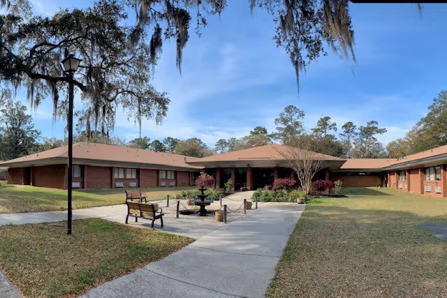 Exterior view of a single-story brick building with a brown roof, surrounded by grass and trees. A concrete walkway leads to the entrance, with benches and a small fountain along the path. The sky is clear with some light clouds.