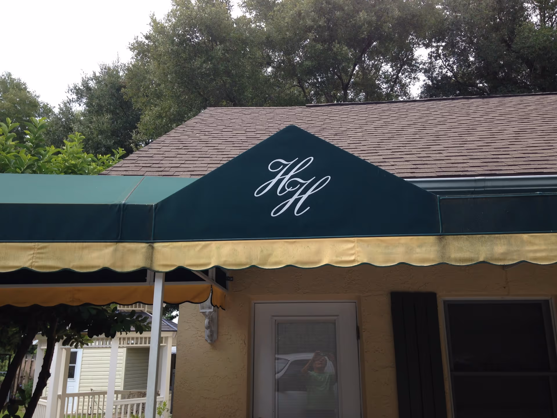 Green and yellow awning with the initials 'HH' in white script above a door on a building with a shingled roof and trees in the background.