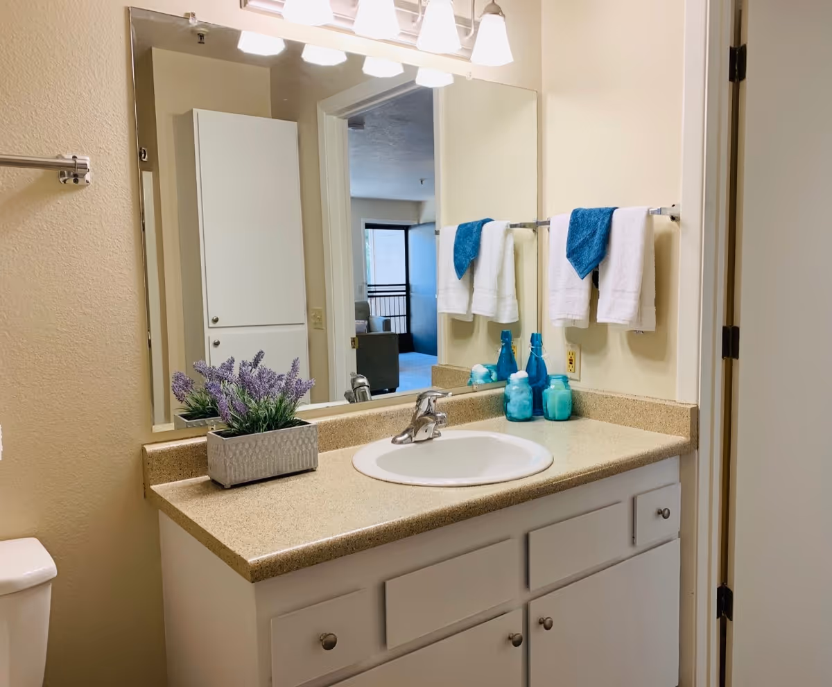 A clean bathroom vanity with a beige countertop, white sink, and silver faucet. Above the sink is a large mirror reflecting a doorway and part of a living area. On the countertop, there is a small rectangular planter with purple flowers and three blue glass containers. Two white towels with blue hand towels are hanging on a towel rack mounted on the wall. The bathroom has light-colored walls and white cabinetry below the sink.