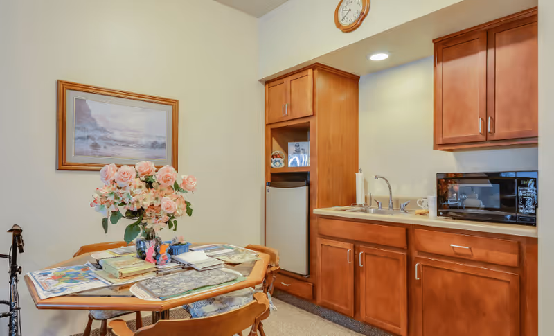 A small kitchen area with wooden cabinets, a sink, a microwave, and a mini refrigerator. In front of the kitchen is a wooden table with four chairs, decorated with a vase of pink flowers and various papers and books on the table. A framed landscape painting hangs on the wall above the table, and a clock is mounted above the cabinets.