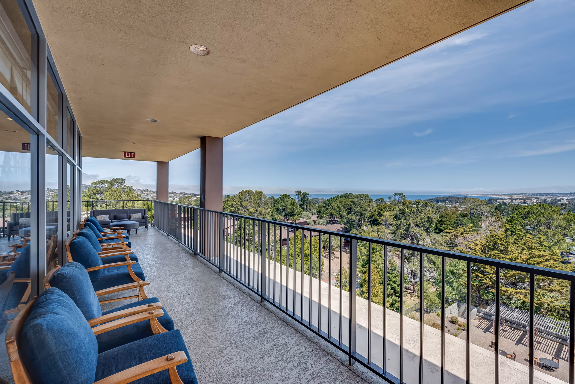 Covered outdoor balcony with blue cushioned wooden chairs lined up along a glass wall on one side and a black metal railing on the other, overlooking a scenic view of trees, buildings, and a distant body of water under a clear blue sky.