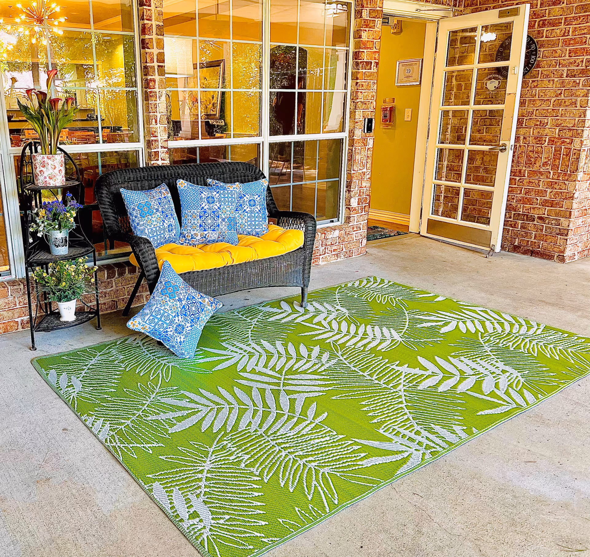 A wicker bench with a yellow cushion and blue patterned pillows sits on a green leaf-patterned rug in front of a brick building entrance.
