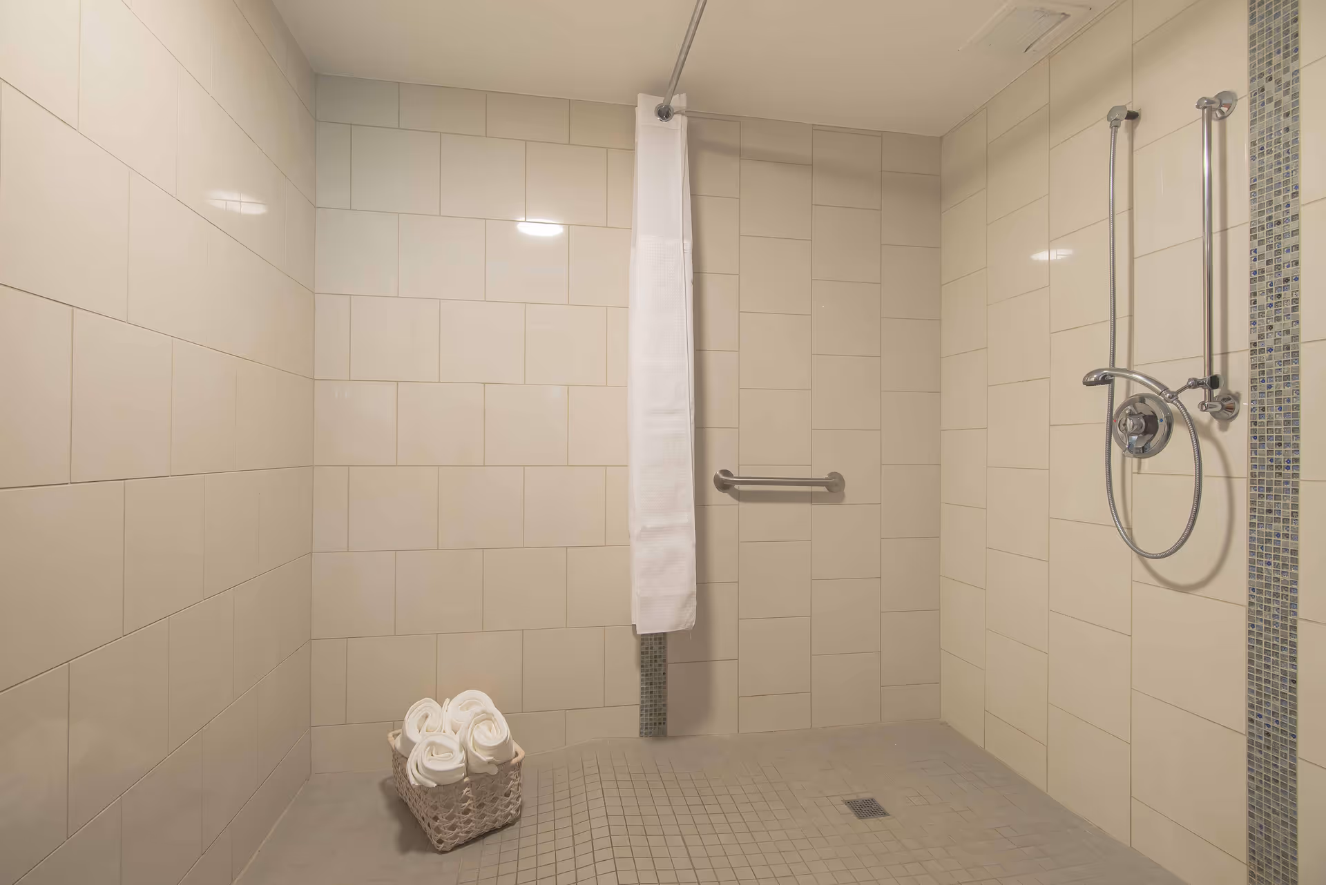 A spacious walk-in shower with beige tiled walls and floor, a white shower curtain partially drawn, a metal grab bar on the wall, a handheld showerhead, and a basket with rolled white towels on the floor.