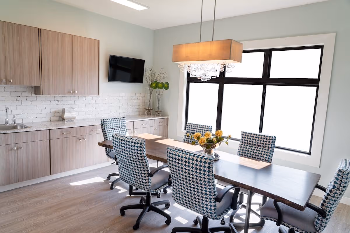 A bright and modern dining area with a wooden table surrounded by six patterned swivel chairs. The room features light wood cabinetry with a white brick backsplash, a wall-mounted flat screen TV, a large window letting in natural light, and a rectangular chandelier with hanging glass orbs above the table. A vase with yellow flowers is placed on the table.