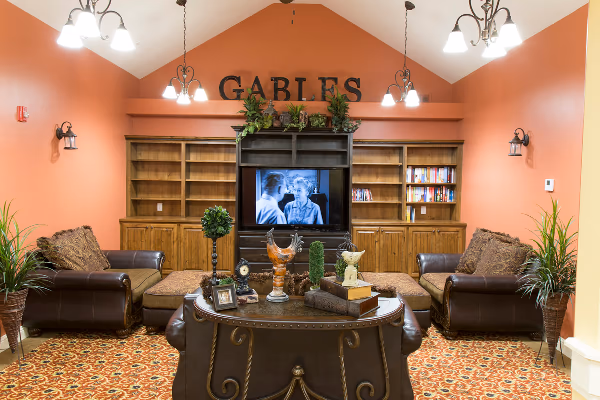 A cozy living room area with two brown leather armchairs and a matching ottoman, a patterned carpet, and a wooden entertainment center with shelves and a TV playing a black-and-white movie. The walls are painted a warm orange color, and there are several decorative plants and ornaments on the table and shelves. Above the entertainment center, large letters spell out 'GABLES'.