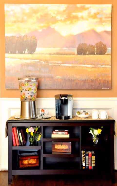 Coffee station with beverage dispensers, cups, flowers, and a landscape painting on the wall.