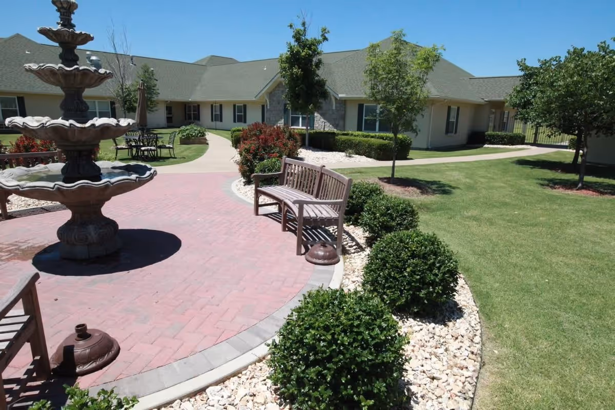Outdoor courtyard area of a senior living facility with a multi-tiered stone fountain, wooden benches, manicured bushes, trees, and a paved walkway leading to a single-story building with a green roof and beige walls under a clear blue sky.