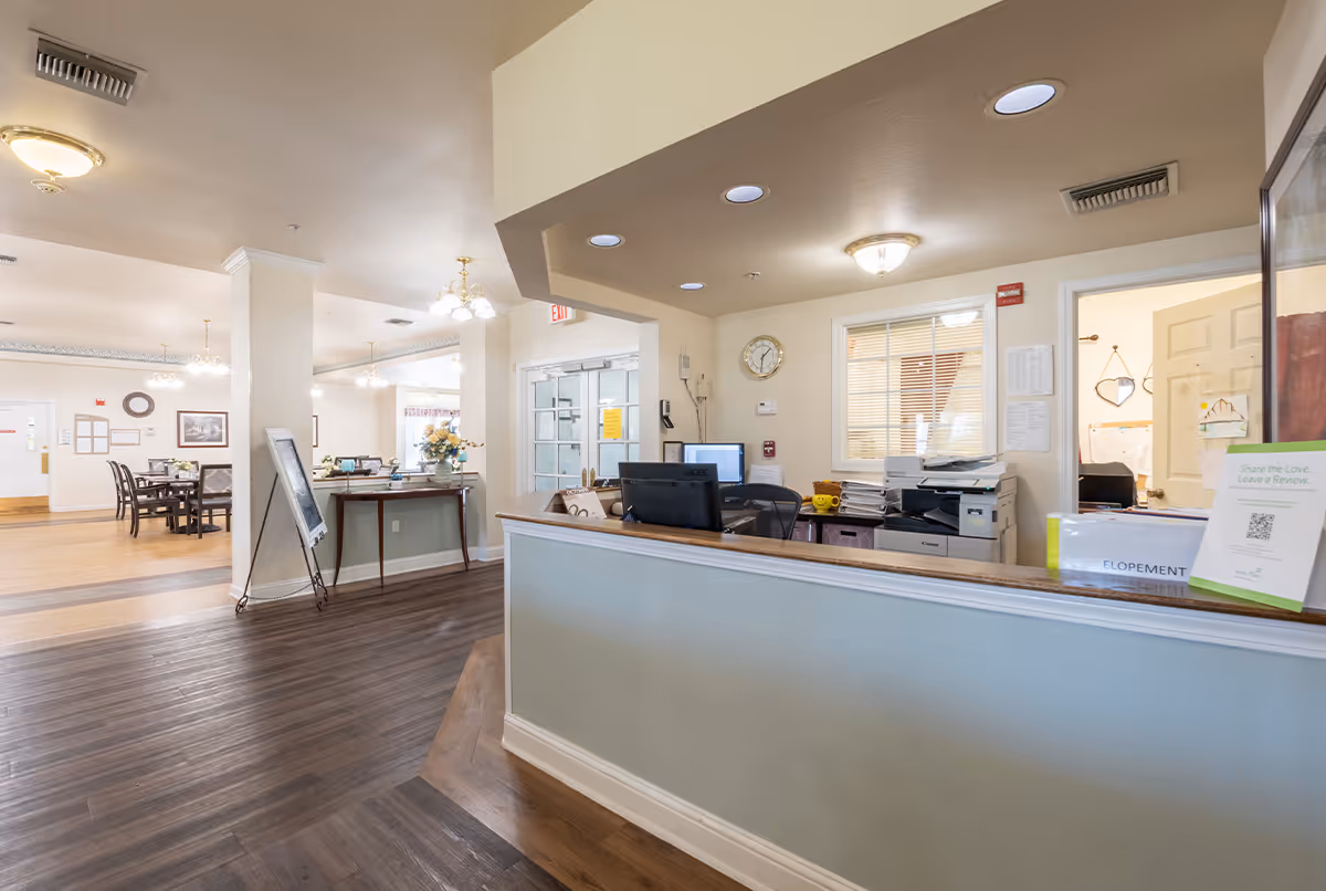 Reception area of a senior living facility with a front desk equipped with computers, a printer, and office supplies. Behind the desk is a window and an open door leading to another room. To the left, there is a dining area with tables and chairs, decorated with flowers and wall art. The space has wood flooring, light-colored walls, and ceiling lights.