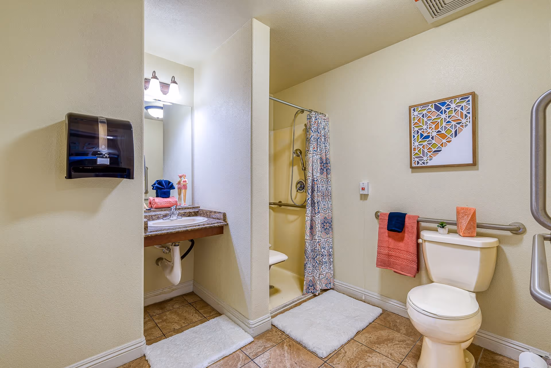 A clean and accessible bathroom with a toilet, a shower with a patterned curtain, a sink with a mirror above it, and grab bars for support. The bathroom has beige walls and tiled floors, with white bath mats on the floor. There are colorful towels and a small decorative plant on the toilet tank, and a framed geometric artwork on the wall.