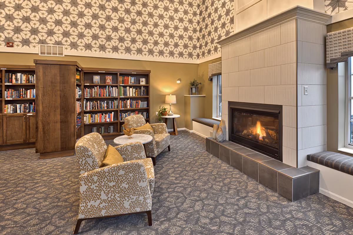 Cozy common room with patterned wallpaper, armchairs around small tables, a fireplace, and bookshelves.
