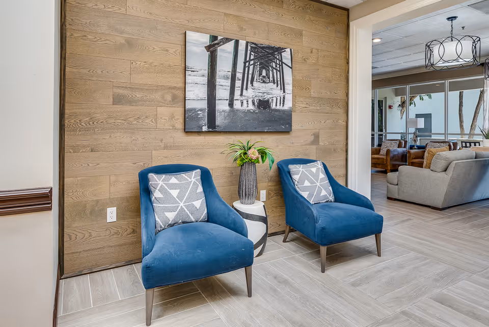 Two blue upholstered chairs with patterned cushions are placed in front of a wooden paneled wall with a black and white photo of a pier above them. A small round table with a vase of flowers is between the chairs. In the background, there is a living area with sofas and large windows.