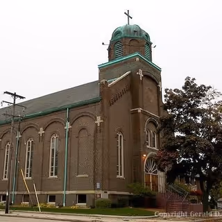 Exterior view of a large brick church building with arched windows and a green dome topped with a cross. There are trees and a sidewalk in front of the building.