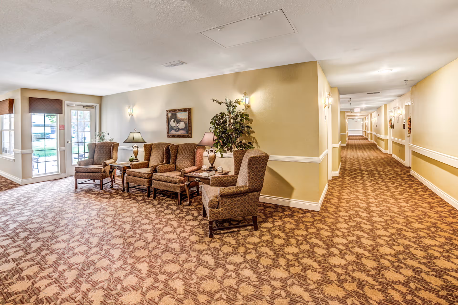Seating area with armchairs, side tables and lamps beside a long carpeted hallway in a senior living facility.