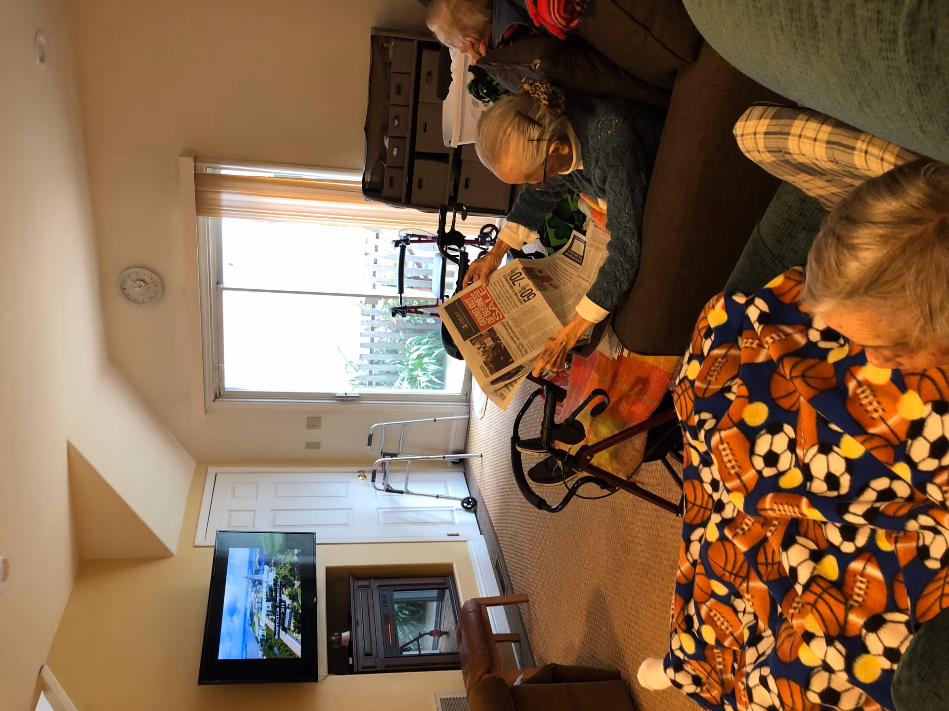 A cozy living room in a senior care home with two elderly women sitting on couches. One woman is reading a newspaper while the other is resting under a blanket with sports ball patterns. The room has a wall-mounted TV, a clock above sliding glass doors, a walker, and storage bins in the background.