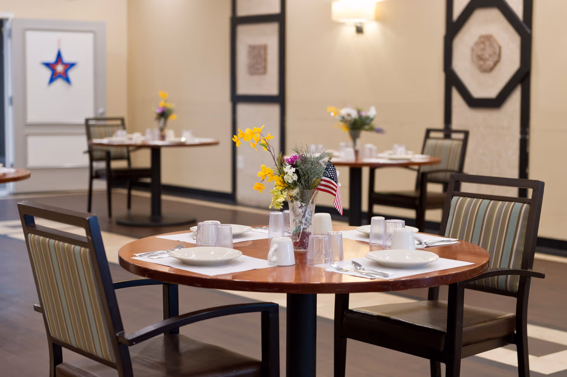 A dining room with round wooden tables set with white plates, cups, and silverware. Each table has a small vase with flowers and an American flag. The room has beige walls with decorative panels and striped cushioned chairs around the tables.