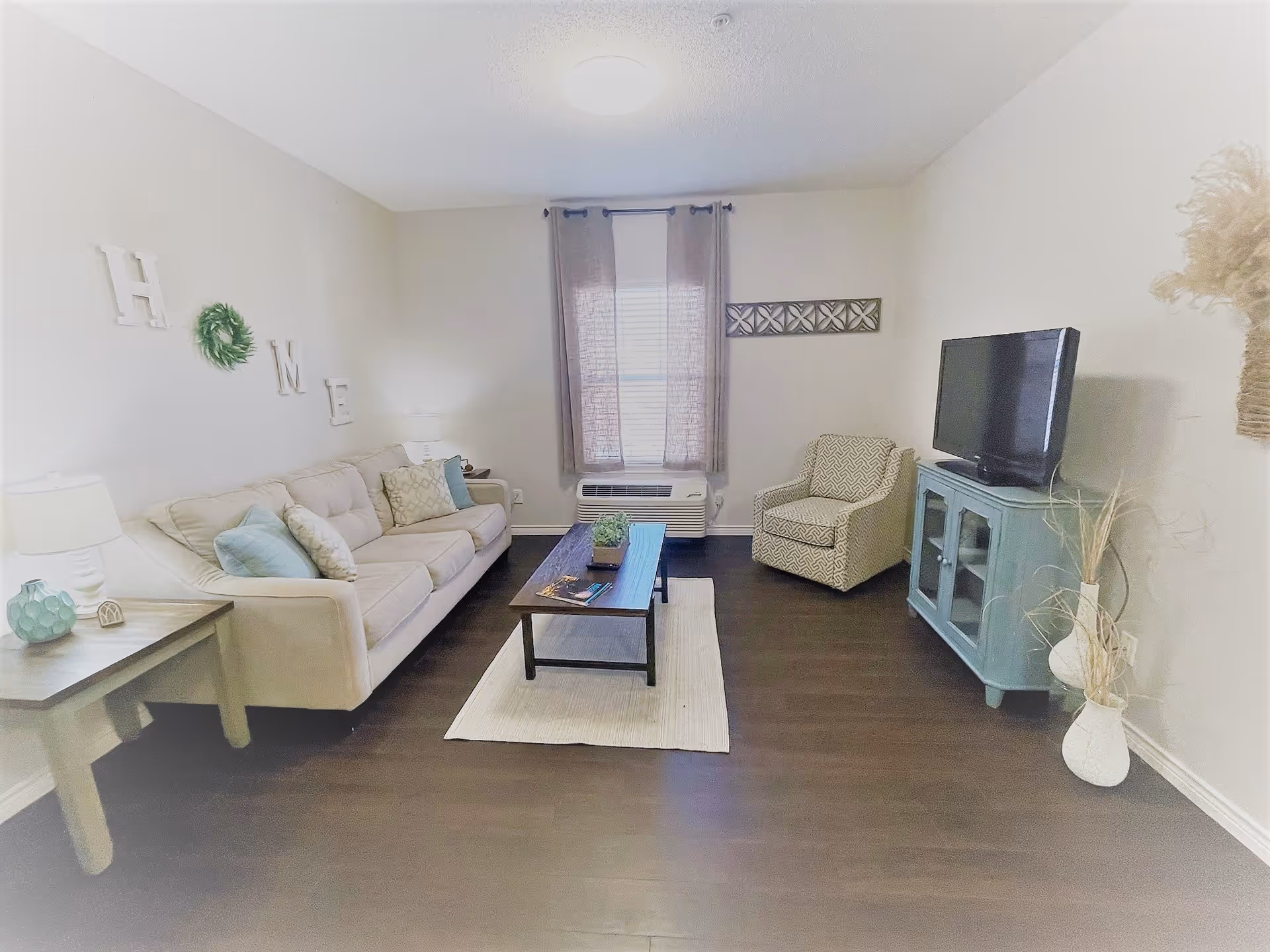 A cozy living room with a beige sofa adorned with cushions, a patterned armchair, a wooden coffee table on a light rug, and a blue TV stand holding a flat-screen television. The walls are light-colored with decorative elements including the word 'HOME' and a small green wreath. A window with sheer curtains is centered on the back wall, and there are two white vases with dried plants on the floor near the TV stand.