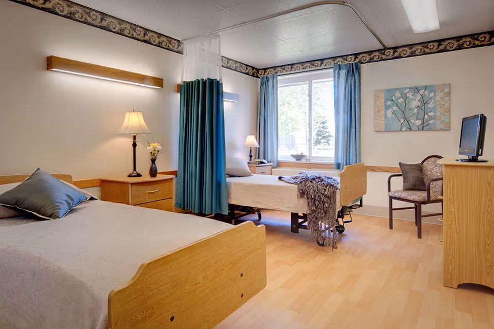 A bright and clean senior living bedroom with two single beds separated by a teal privacy curtain. Each bed has a pillow and a light-colored bedspread. There are two bedside tables with lamps and a vase with flowers. A window with blue curtains lets in natural light. A wooden dresser with a small TV and a cushioned chair with a pillow are also visible. The room has light wood flooring and neutral-colored walls with a decorative border near the ceiling.