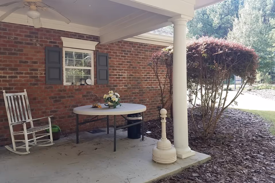 Covered outdoor patio area with a white rocking chair, a round table with a flower arrangement, a ceiling fan, and a brick wall with a window. There is a white column supporting the roof and trimmed bushes nearby.