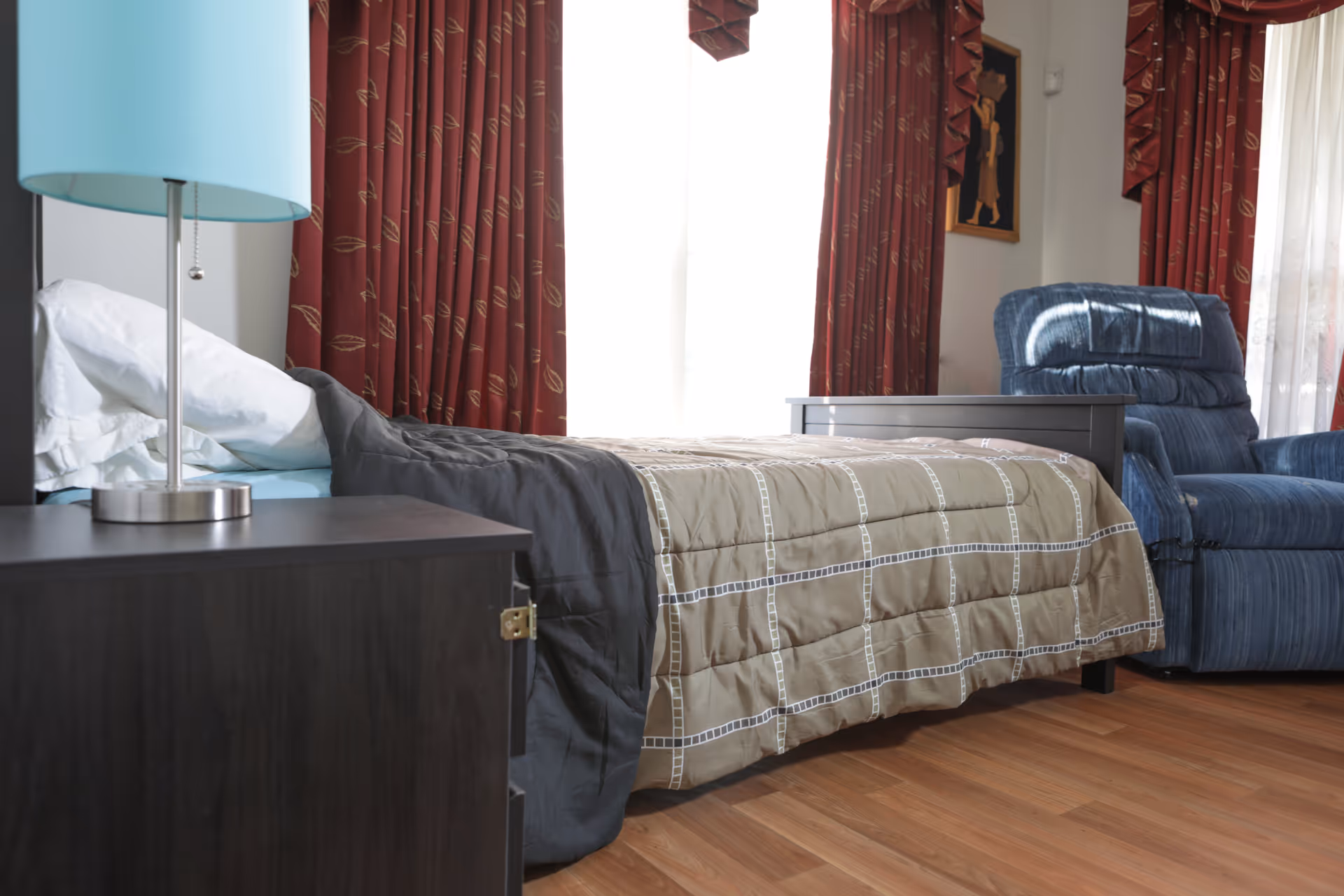 A bedroom with a made bed, nightstand and lamp, and a blue recliner in front of red curtains.