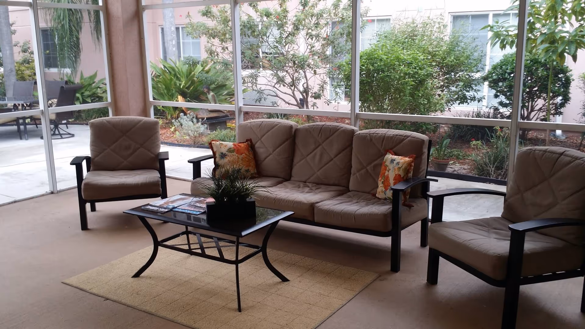 A screened-in patio area with a beige cushioned sofa and two matching armchairs around a black metal coffee table with a plant and magazines on it. Outside the screened area, there are green bushes and trees with a pink building in the background.