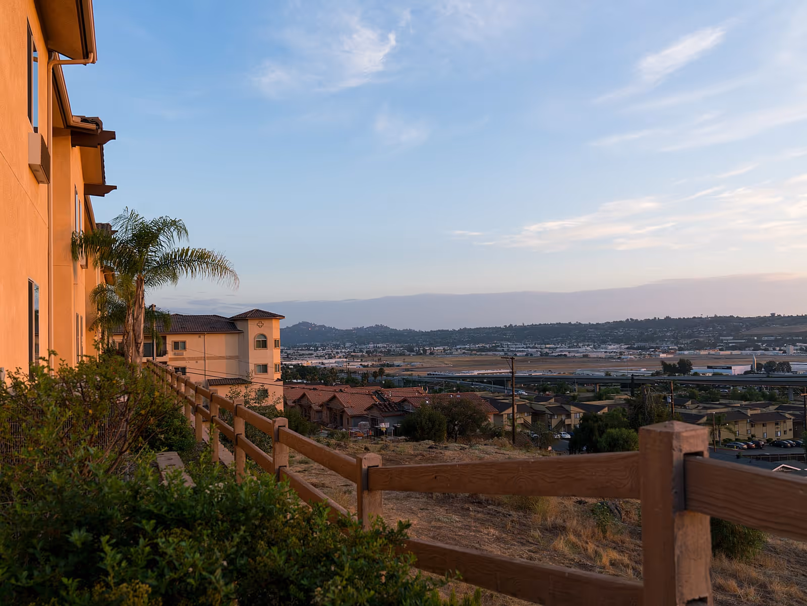 View from a hillside near a senior living facility showing a wooden fence, bushes, palm trees, and buildings with a cityscape and hills in the background under a partly cloudy sky.