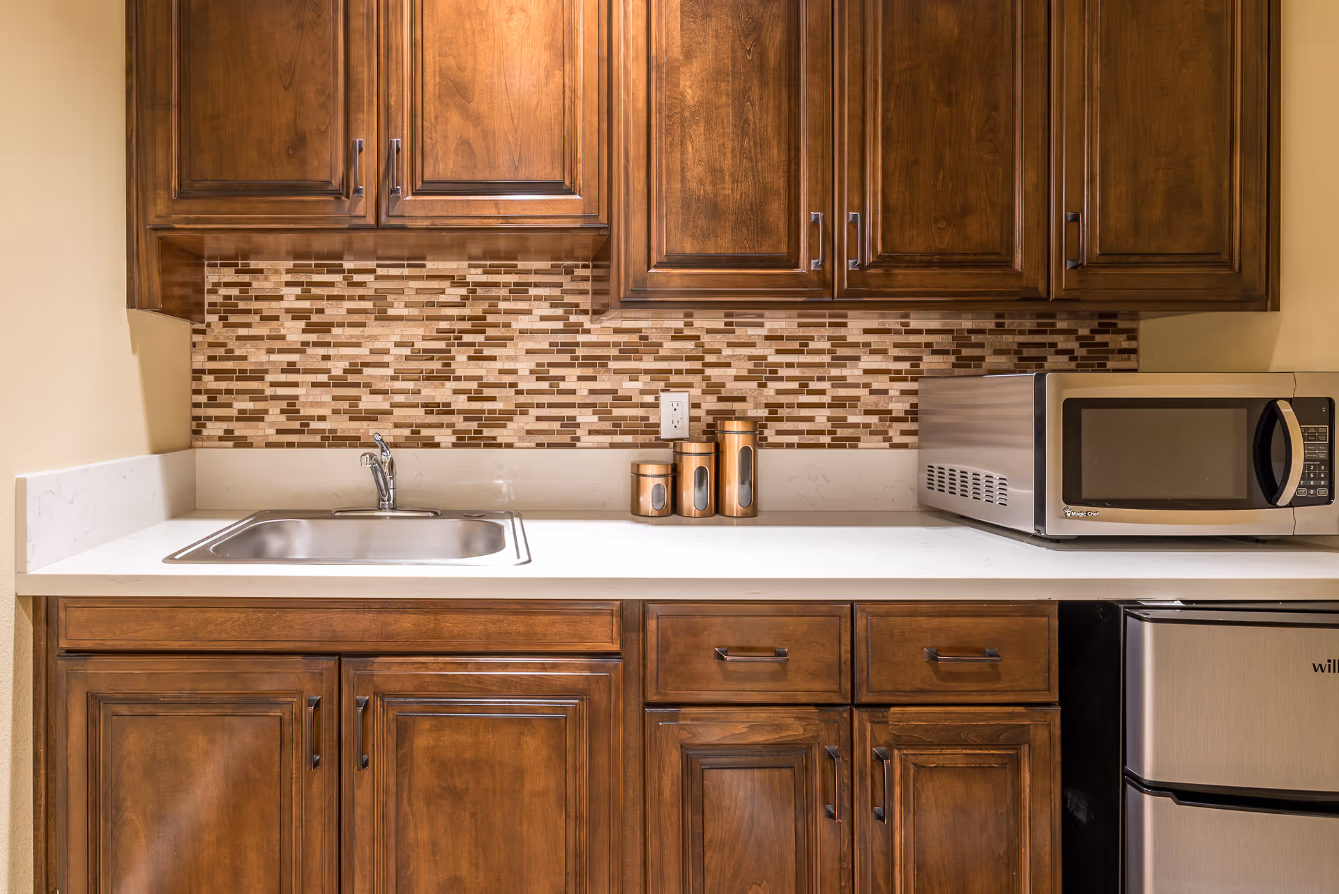 A kitchen area with wooden cabinets, a stainless steel sink, a white countertop, a mosaic tile backsplash in shades of brown and beige, three copper canisters, a stainless steel microwave, and a small stainless steel refrigerator.