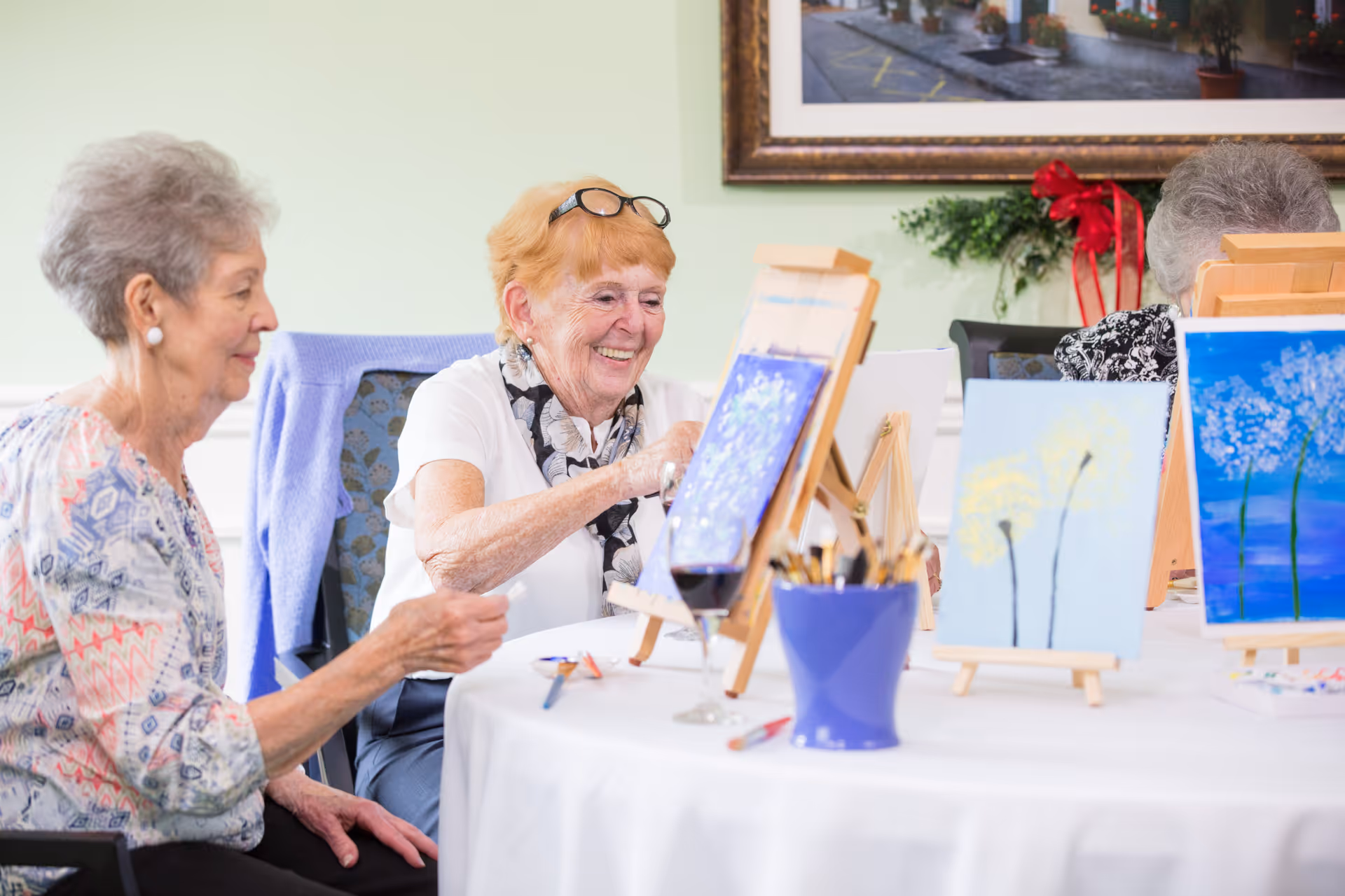 Two elderly women sitting at a round table engaged in painting on canvases set on easels, with paintbrushes and art supplies on the table, in a well-lit room with a framed picture on the wall behind them.