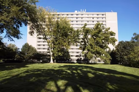 Multi-story apartment building viewed from a grassy lawn with trees under a clear blue sky.