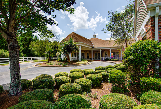 Exterior view of a senior living facility named Oaks at Maple Ridge, showing a driveway with neatly trimmed bushes and trees, a white fence, and a building with a covered entrance under a partly cloudy sky.