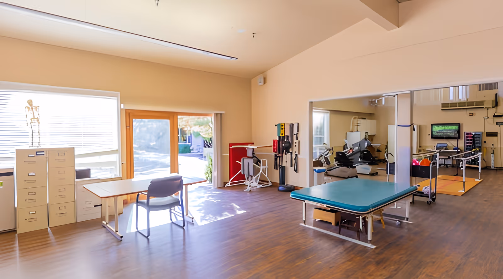 A spacious physical therapy room with wooden flooring, a treatment table, exercise equipment, a desk with a chair, filing cabinets, and large windows letting in natural light. There is a large mirror on one wall and a door leading outside.