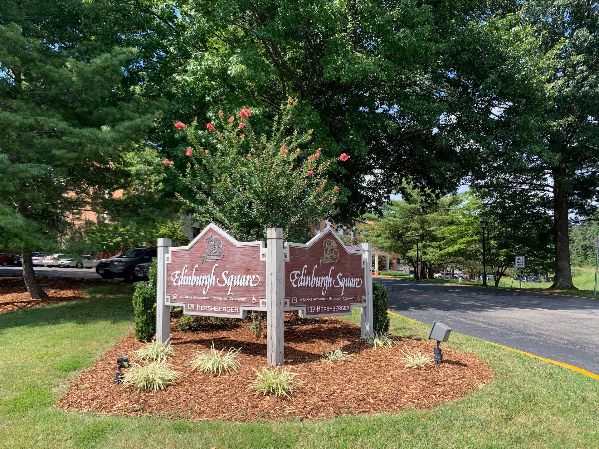A wooden sign for Edinburgh Square, a caring affordable retirement community, located at 129 Hershberger. The sign is surrounded by mulch and small plants, with trees and a parking lot in the background under a clear sky.