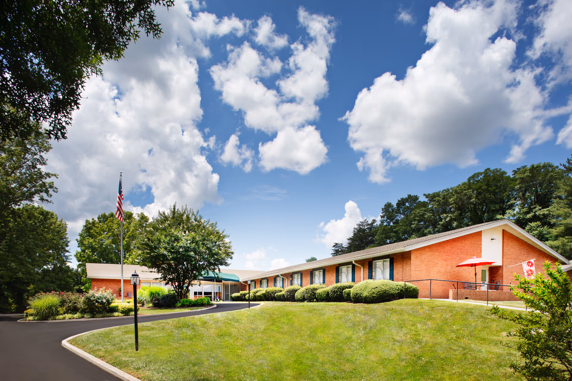 Single-story brick nursing home building with a manicured lawn, American flag, and a partly cloudy sky.