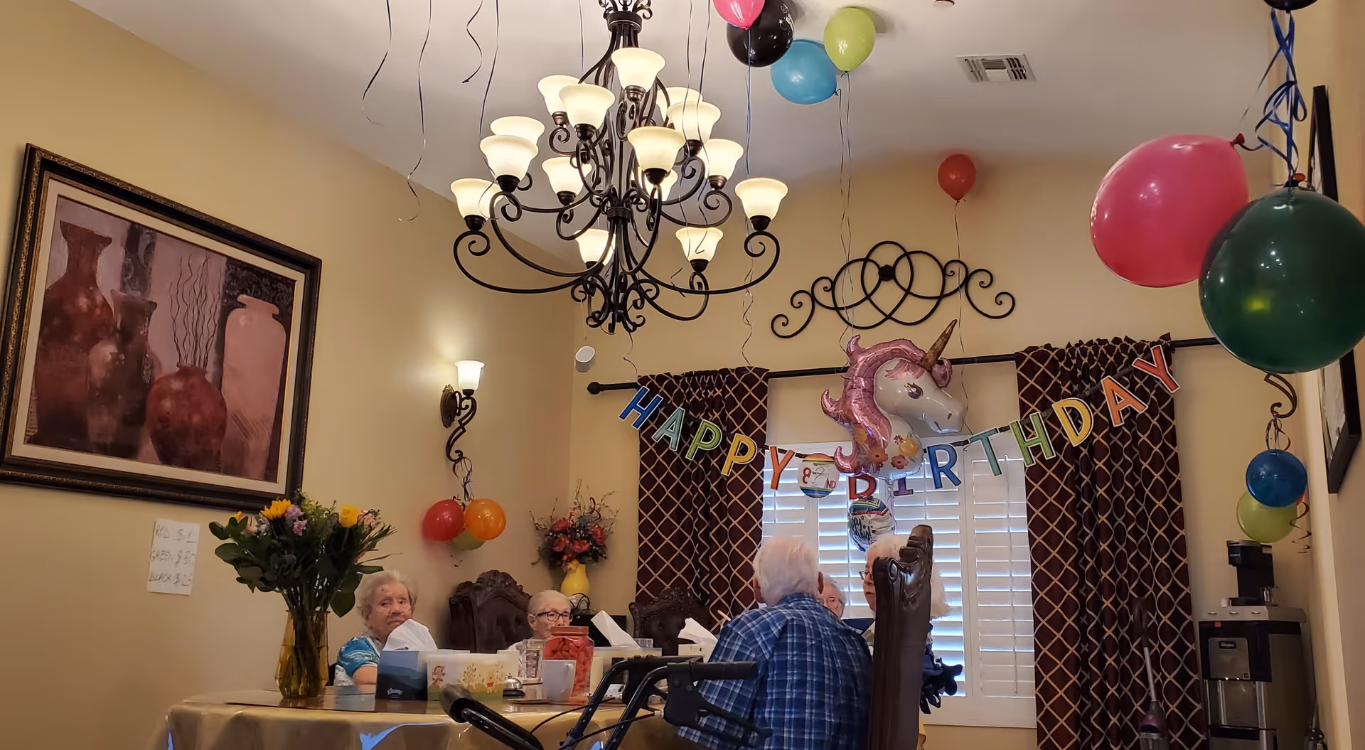 Decorated dining room with elderly residents seated at a table under a chandelier and a 'Happy Birthday' banner with balloons.