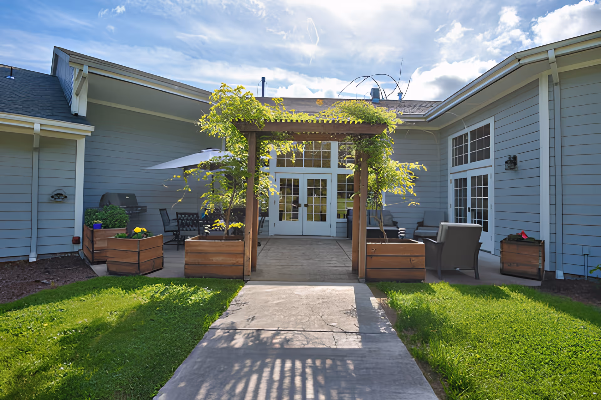 Entrance courtyard of a light-blue assisted living building with a wooden pergola, planters, patio furniture, and double glass doors.