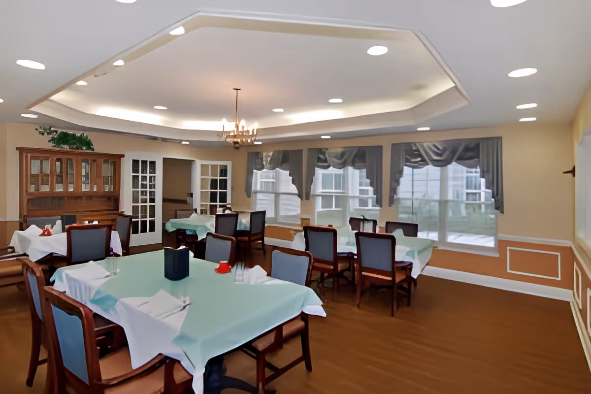 A dining room with several tables covered with light blue tablecloths and white overlays, each surrounded by wooden chairs with blue cushions. The room features large windows with dark valances, a wooden cabinet with glass doors, and a chandelier hanging from a recessed ceiling with soft lighting.