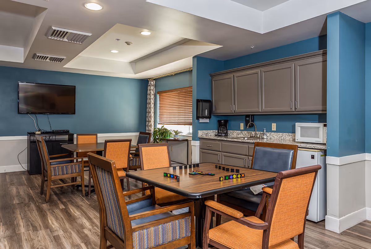 Community dining/activity room with tables and chairs, a wall-mounted TV, and a kitchenette with cabinets and microwave.
