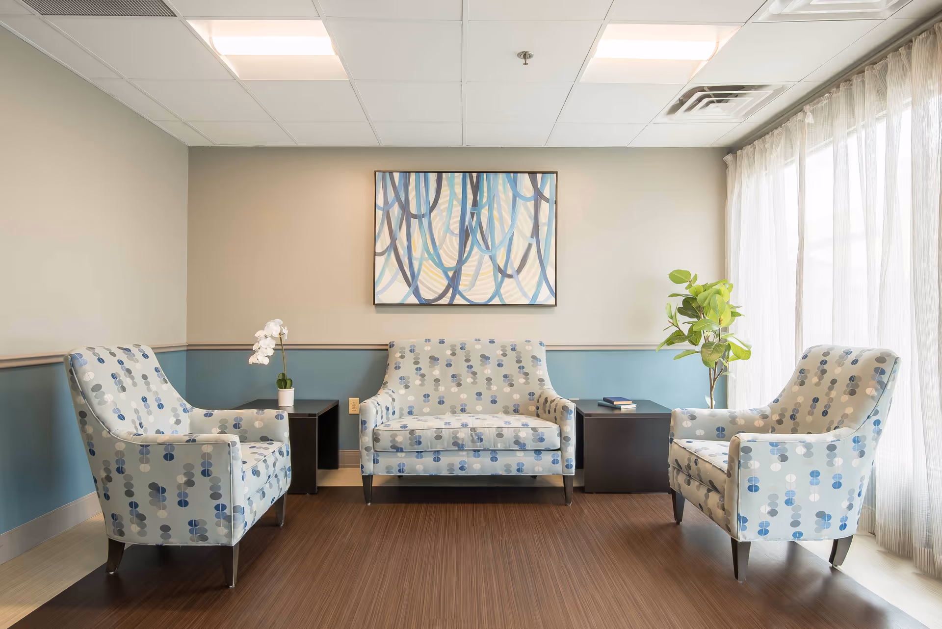 A bright and cozy sitting area with two patterned armchairs and a matching loveseat arranged around two dark wooden side tables. One side table has a small white orchid plant, and the other has a book and a green leafy plant. A large abstract painting with blue and beige tones hangs on the wall above the loveseat. Sheer white curtains cover the window, allowing natural light to fill the room.